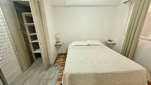 Un dormitorio con cama, mesita de noche, lámpara, armario y una puerta que da a otra habitación.