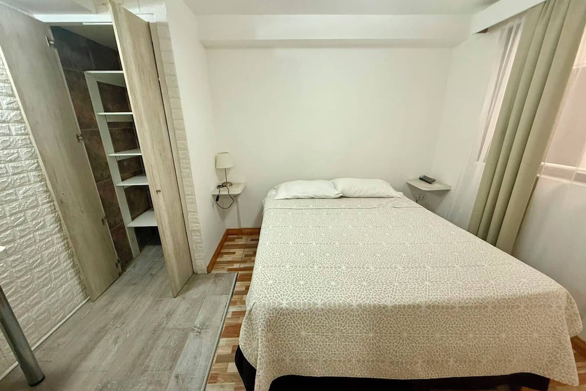 A bedroom with a bed, a nightstand, a lamp, a wardrobe, and a door leading to another room.