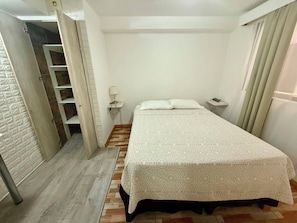 Un dormitorio con cama, mesita de noche, lámpara, armario y una puerta que da a otra habitación.