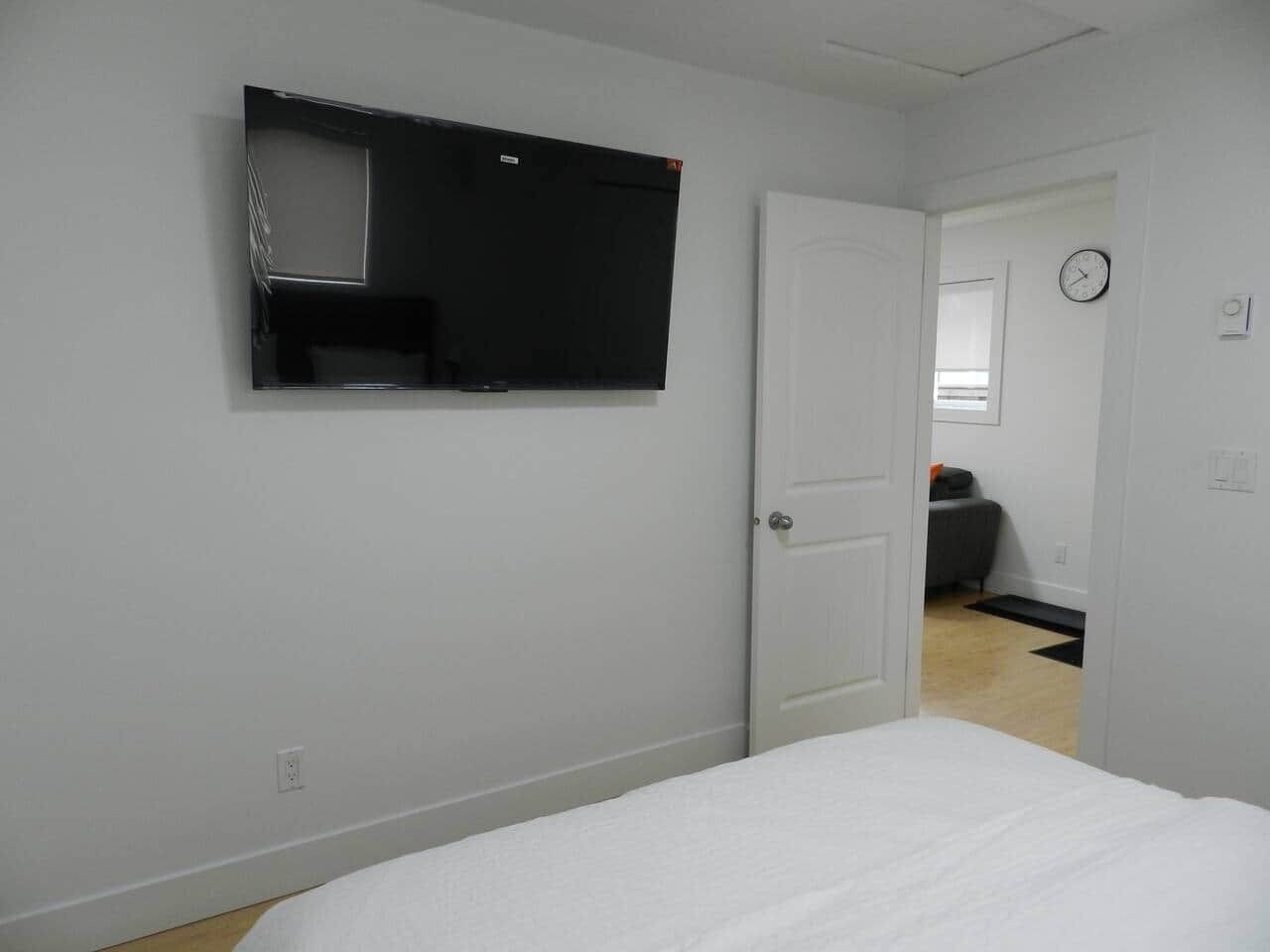 Een slaapkamer met een bed, een deur, een aan de muur bevestigde televisie en een klok.