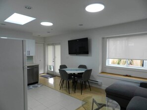A modern kitchen with white cabinets, a dining table with chairs, a flat-screen TV mounted on the wall, and a window with a view.