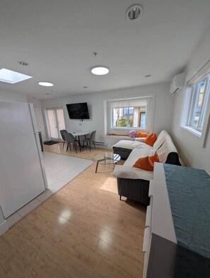 A modern, well-lit room with a sofa, a dining table with chairs, a TV mounted on the wall, and a window providing natural light. - Modern PNE Laneway Home – Quiet Retreat Near Hastings Park (Vancouver)