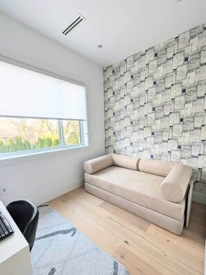 Ein Zimmer mit Fenster, einem beigen Sofa, einem Schreibtisch mit Computer und einer Wand mit Muster.