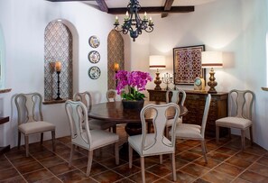 Een eetruimte met een ronde tafel, zes gestoffeerde stoelen, een kroonluchter, decoratieve wandpanelen en een klok.
