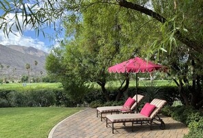 Een terras met twee lounge stoelen onder een rode parasol, omgeven door groen en uitzicht op een berg.
