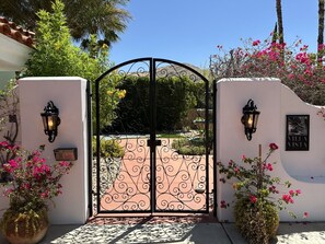 A gated entrance with a wrought iron gate, a wall-mounted sign, and a potted plant with pink flowers. - Indian Canyons Golf Resort Fairway-Front Home Pool & Spa + Golf & Mountain Views (Palm Springs)