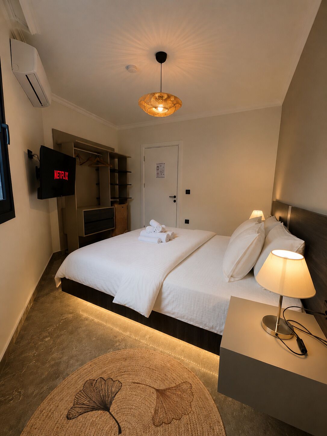 A hotel room with a bed, a TV mounted on the wall, a door, a bedside table with a lamp, and a decorative rug.