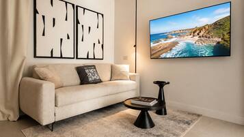 Un soggiorno con un divano color crema, un tavolino da caffè, un quadro incorniciato e una TV a schermo piatto che mostra una veduta costiera.
