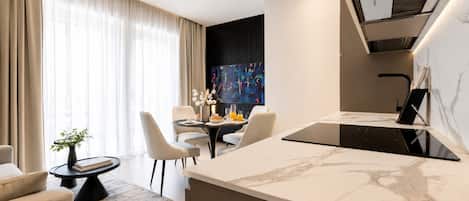 A modern hotel room with a marble kitchen island, a dining area with a table and chairs, a large window with curtains, and a wall-mounted TV.