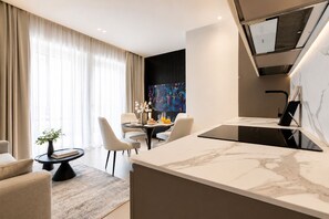 Una habitación de hotel moderna con una isla de cocina de mármol, un comedor con mesa y sillas, un ventanal con cortinas y un televisor montado en la pared.