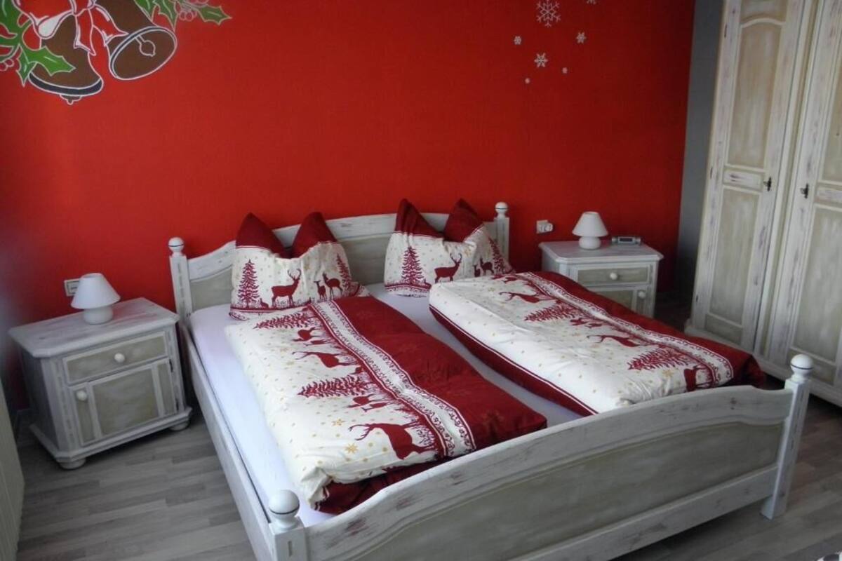 Una cama con estructura blanca y ropa de cama roja, una mesita de noche con lámpara y un armario en una habitación con paredes rojas.