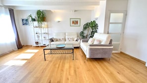 Una sala de estar con una mesa de centro de vidrio, dos sofás blancos, piso de madera y una planta grande.