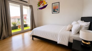 Un dormitorio con cama, mesita de noche, lámpara, ventana con cortinas e imagen en un marco en la pared.