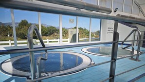 An indoor swimming pool with two separate water areas, surrounded by a metal railing, and a view of the outdoors through large windows.