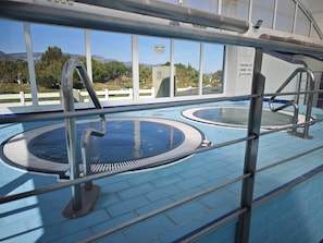Une piscine intérieure dotée de deux espaces aquatiques distincts, entourée d’une rambarde métallique, et offrant une vue sur l’extérieur grâce à de grandes fenêtres.
