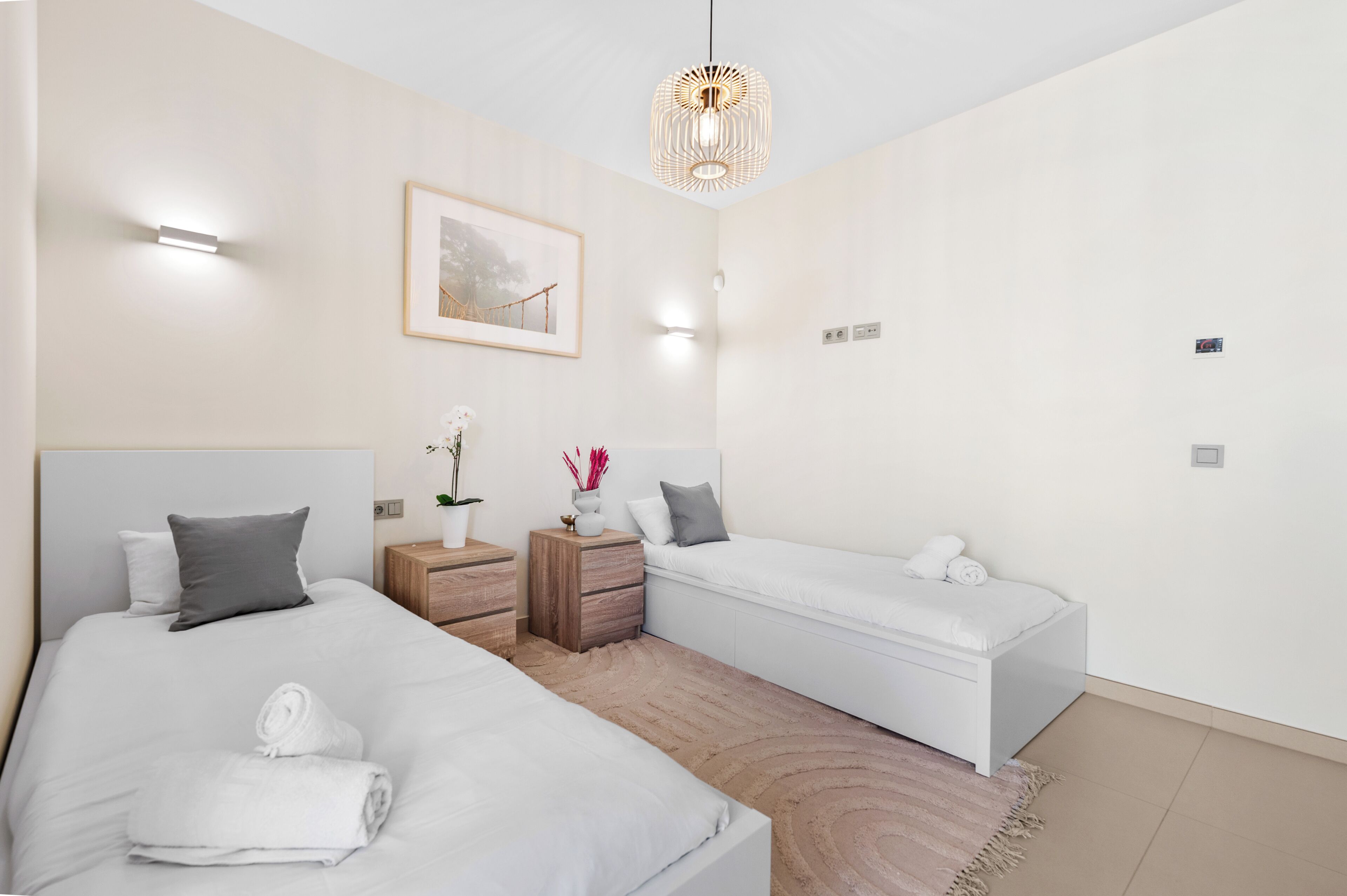 A modern hotel room with two beds, a wooden nightstand, a small table with a vase, and a hanging light fixture.