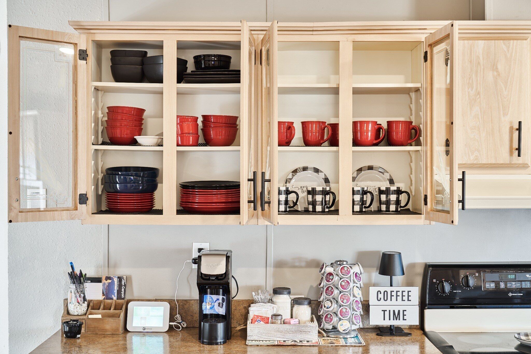 A kitchen with open wooden cabinets containing red and black dishes, a coffee station with a coffee maker, and a sign that reads 'COFFEE TIME'.