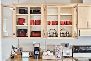 Una cocina con alacenas de madera abiertas que contienen vajilla roja y negra, una estación de café con cafetera, y un cartel que dice 'COFFEE TIME'.