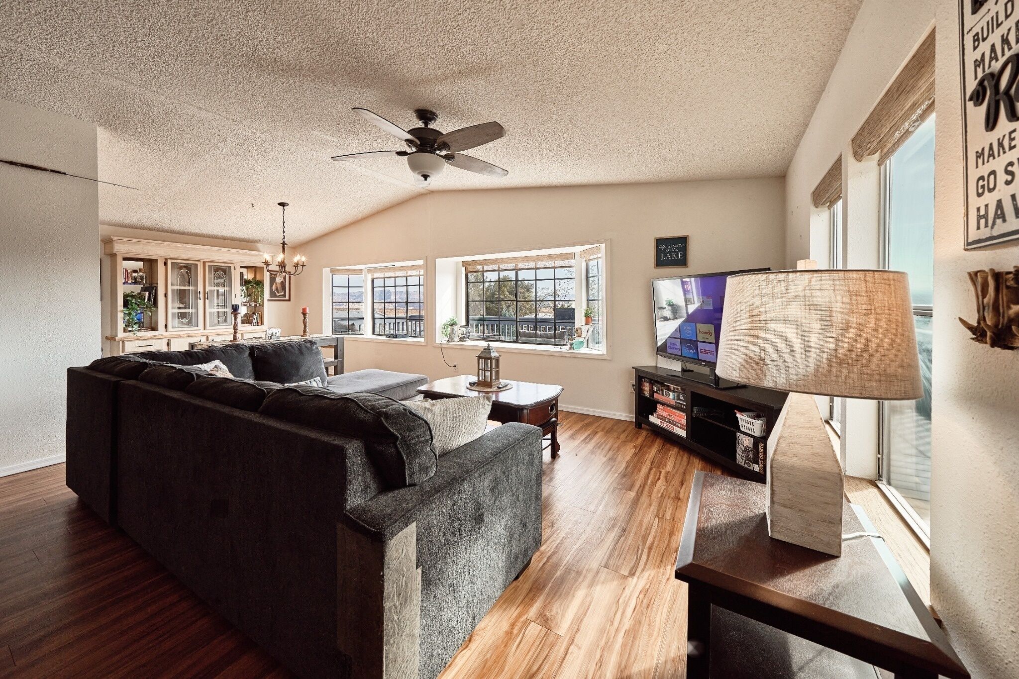 A living room with a dark sofa, wooden flooring, a ceiling fan, a television, and a bookshelf.