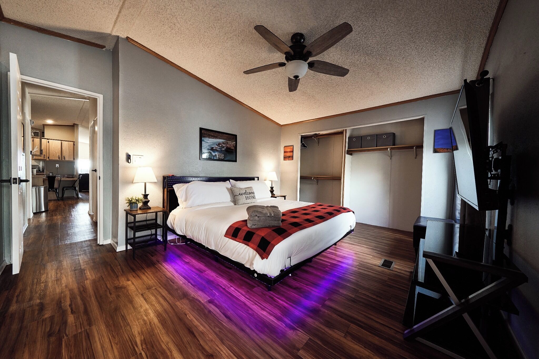 A bedroom with a bed, a ceiling fan, a nightstand, a lamp, a painting, and a view of the kitchen.