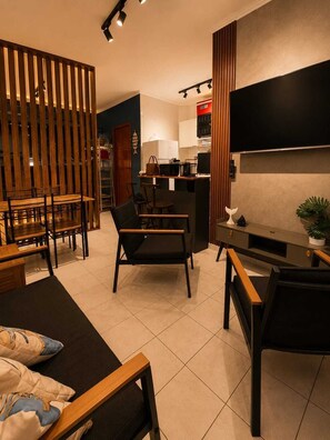 A modern living room with a sofa, chairs, a TV, a kitchen area, and a wooden partition. - Kitnet na Ocian - 500 metros do mar, Ar condicionado  e garagem (Praia Grande)