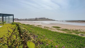 Eine Küstenlandschaft mit einem modernen Gebäude, einem Strand und einer entfernten Hügelkette.
