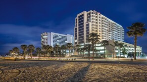 Un hôtel situé en bord de mer, avec plusieurs étages, des palmiers et une plage de sable.