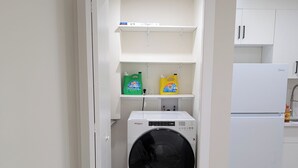 A laundry room with a front-loading washer and dryer, a shelf with cleaning supplies, and a white refrigerator. - New Cozy 2 Bedrooms Basement Apartment (Calgary)