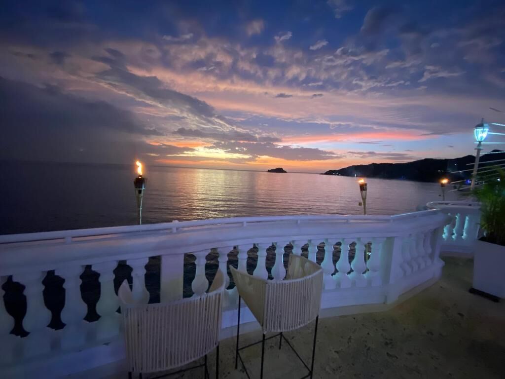 Ein Balkon mit Blick auf das Meer bei Sonnenuntergang, ein weißes Geländer und zwei Stühle.