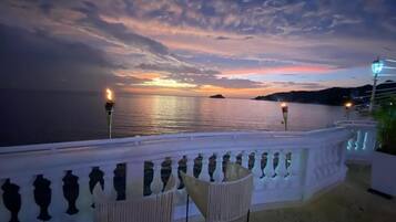 Ein Balkon mit Blick auf das Meer bei Sonnenuntergang, ein weißes Geländer und zwei Stühle.