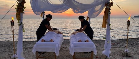 A beachside massage setup with two massage tables under a canopy, surrounded by candles and flowers.