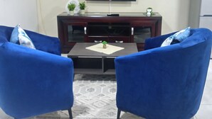 A living room with two blue armchairs, a coffee table, a flat-screen TV, a small plant, and a refrigerator. - Sydney's Guest house
 (Johannesburg South)