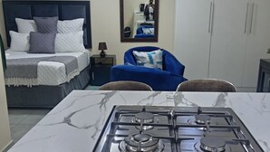 A kitchen with a stove, a bed with pillows, a chair, and a mirror on the wall. - Sydney's Guest house
 (Johannesburg South)