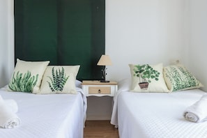 Ein Zimmer mit zwei Betten, jedes mit einem Kissen mit Pflanzenmuster, einem Nachttisch mit Lampe und einer kleinen Schublade.