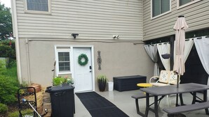 Un patio con una mesa, sillas, un cesto de basura negro, una planta en maceta y una puerta decorada con un adorno.