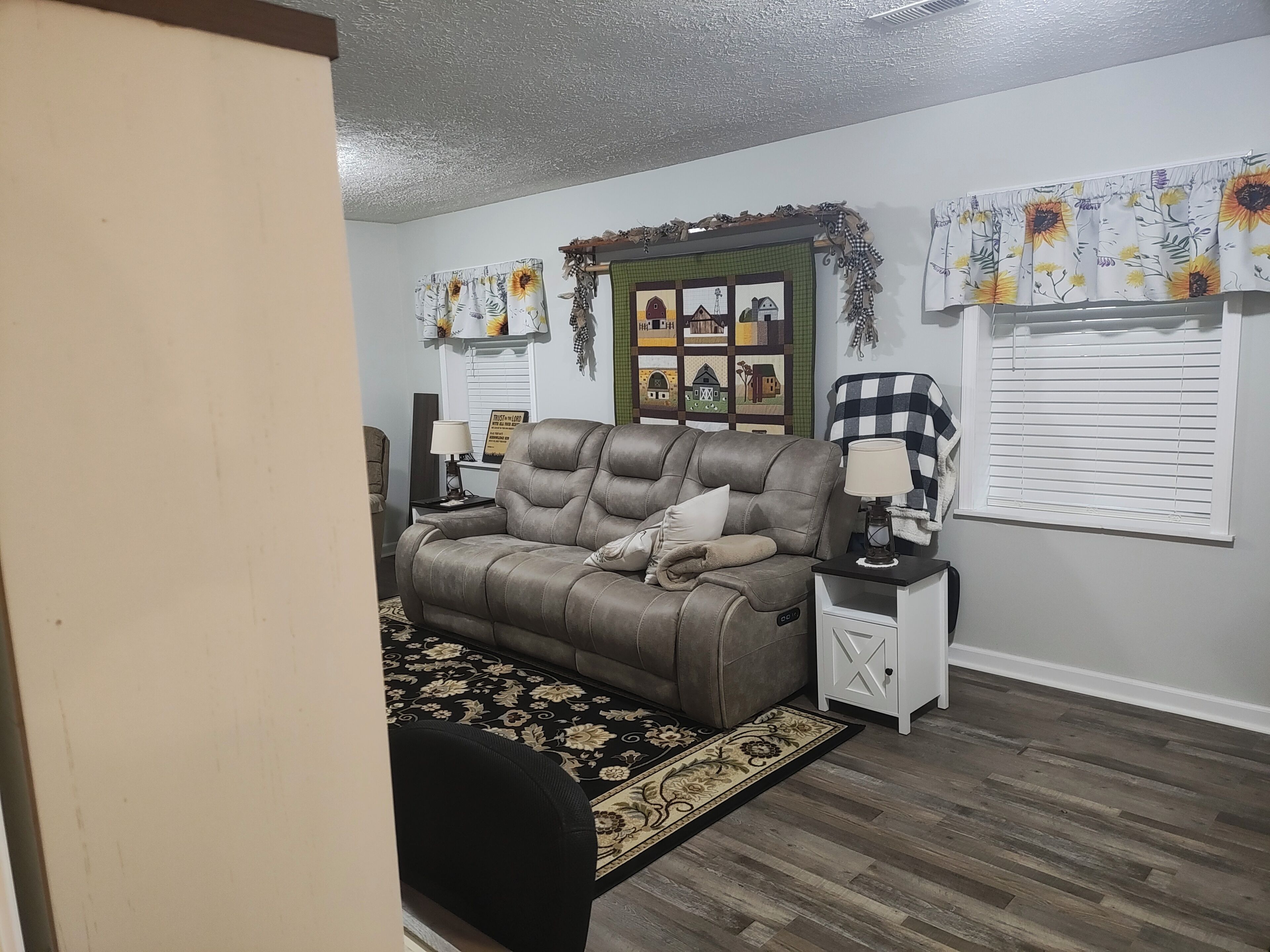 A living room with a brown leather sofa, a black and beige floral rug, a white side table with a lamp, and a wall decorated with sunflower prints and a green-framed picture shelf.