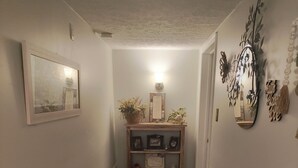 Un pasillo con piso de madera, un tapete oscuro con diseño, una pequeña consola de madera con objetos decorativos, un cuadro enmarcado y un espejo montado en la pared.