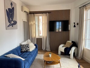 Un salon avec un canapé bleu, une table basse en bois, un fauteuil blanc, un téléviseur fixé au mur et une fenêtre avec des rideaux.