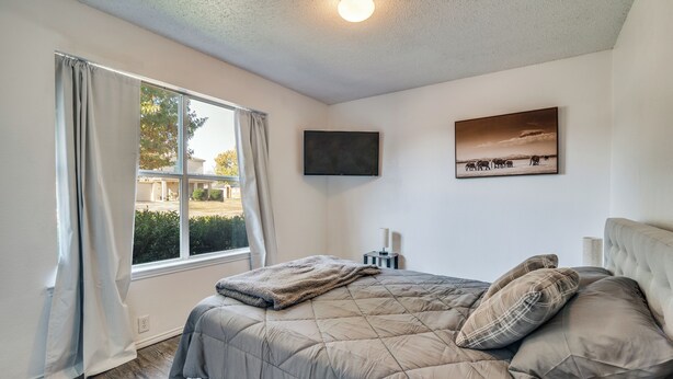Ein Schlafzimmer mit einem Bett, einem Fenster mit Vorhängen, einem Flachbildfernseher und einem Bild an der Wand.