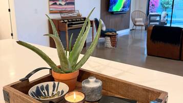 Un plateau en bois sur lequel on a posé un pot d’aloès, une bougie, un bol et un livre intitulé « GUÉRIR ET NOURRIR L’AMOUR », dans un salon moderne avec une télévision et des œuvres d’art accrochées au mur.