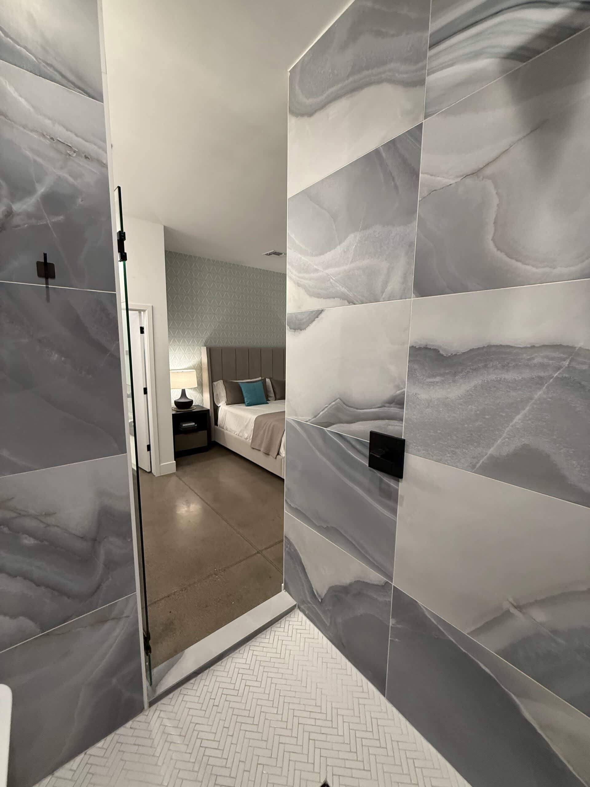 Une chambre d’hôtel avec un parquet à motifs en chevrons, un lit avec une tête de lit, une table de chevet et un mur au motif rappelant le marbre.