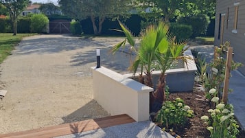 Un camino de entrada residencial con un sendero de grava, una jardinera de concreto con plantas y una terraza de madera.