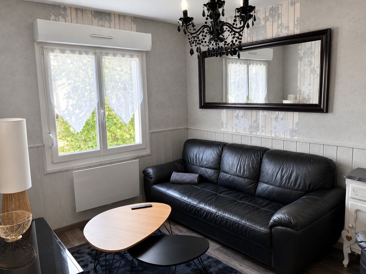 A room with a black leather sofa, a wooden coffee table, a small round table, a lamp, a mirror, and a window with a view of greenery.