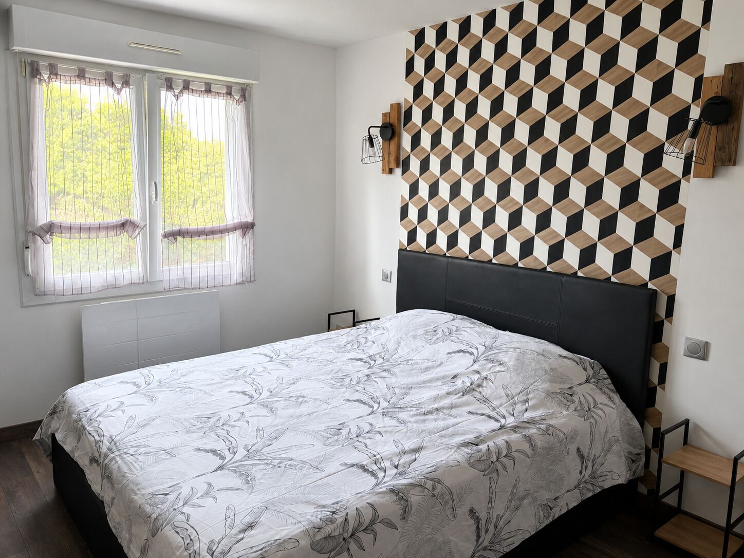 A bedroom with a bed, a window with curtains, a geometric wall pattern, and a headboard.
