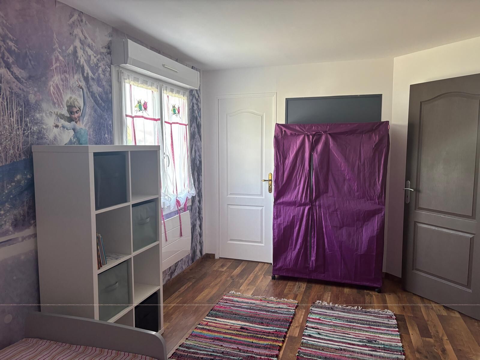 A room with a white bookshelf, a purple wardrobe, a door, and a colorful rug.