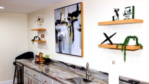 Uma cozinha com bancada de mármore, pia de aço inoxidável e armários de madeira. Há objetos decorativos nas prateleiras e um quadro na parede.