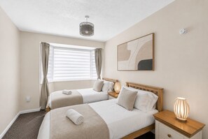 Et hotelværelse med to senge, et vindue med gardiner, en loftlampe, et sengebord med en lampe og et ophængt abstrakt billede på væggen.