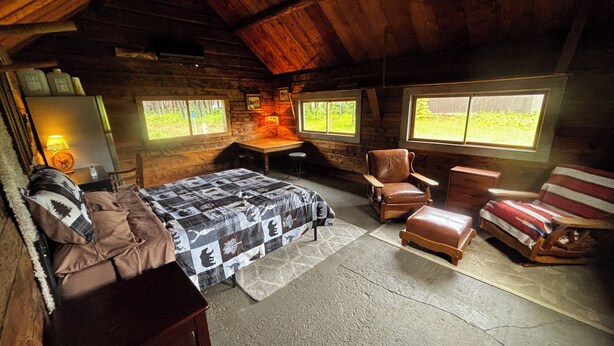 Eine Hütte mit Bett, Schreibtisch, Stuhl und einem Fenster mit Blick auf Bäume.