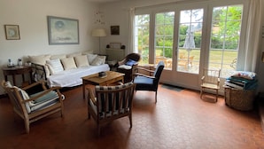 Een gezellige woonkamer met een bank, stoelen, een tafel en uitzicht op een tuin door grote ramen.
