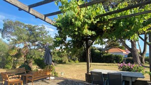 Een terras met stenen vloer, houten meubilair, een pergola begroeid met wijnranken en uitzicht op de zee.